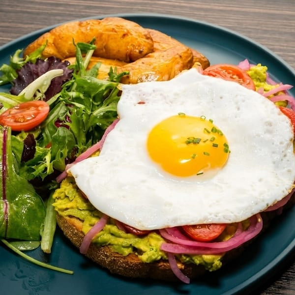 Avocado toast with a fried egg, pickled onions, cherry tomatoes, side salad, and roasted potatoes.
