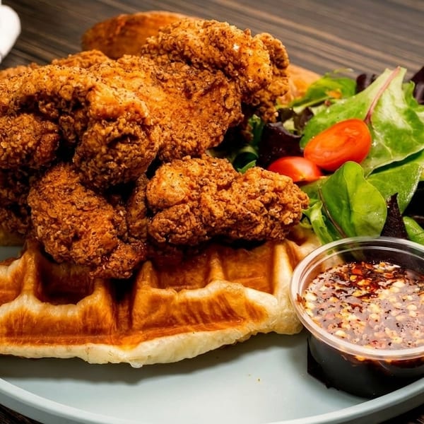 Crispy fried chicken pieces on a waffle with a side salad and chili sauce on a light blue plate.
