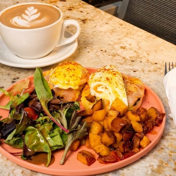 Eggs Benedict with roasted potatoes, a side salad, and a latte on a granite countertop.